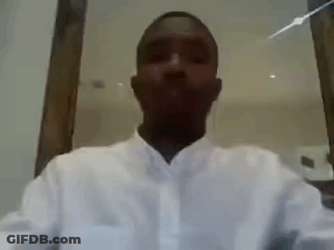 A Man In A White Shirt Is Sitting In Front Of A Mirror And Looking At The Camera . GIF