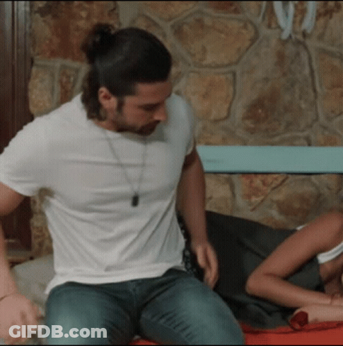 A Man In A White Shirt Is Sitting On A Bed GIF