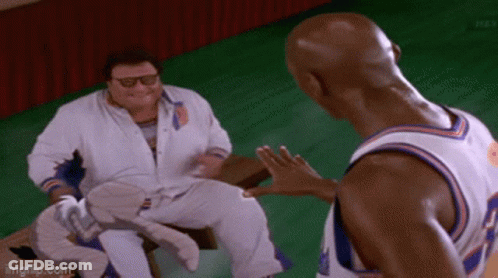 A Man In A White Shirt Is Sitting On A Bench Talking To Another Man In A Basketball Uniform . GIF