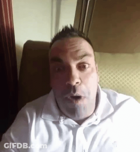 A Man In A White Shirt Is Sitting On A Couch And Making A Surprised Face . GIF