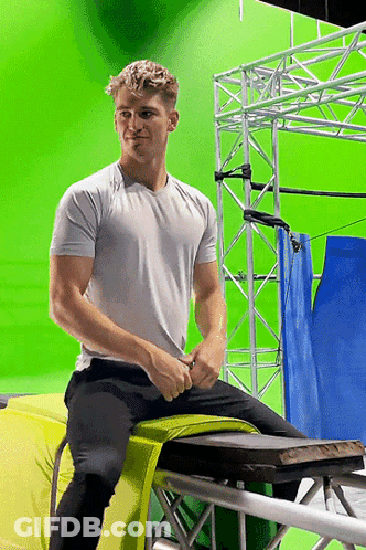 A Man In A White Shirt Is Sitting On A Yellow Cushion In Front Of A Green Screen GIF