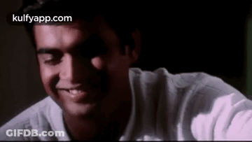 A Man In A White Shirt Is Smiling With His Eyes Closed In A Dark Room . GIF