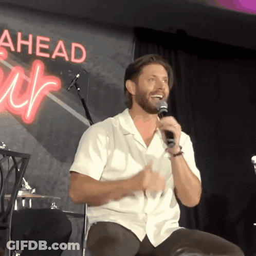 A Man In A White Shirt Is Speaking Into A Microphone In Front Of A Sign That Says Ahead GIF