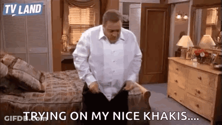 A Man In A White Shirt Is Standing In A Bedroom . GIF
