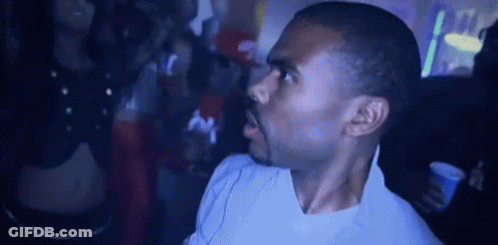 A Man In A White Shirt Is Standing In A Dark Room With A Crowd Behind Him . GIF