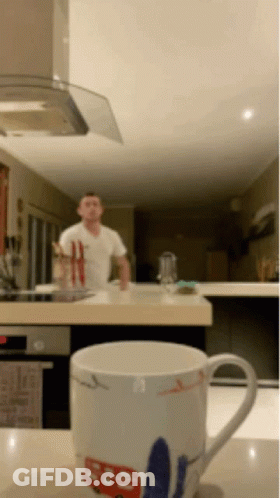 A Man In A White Shirt Is Standing In A Kitchen Behind A Mug That Says ' Sd ' On It GIF