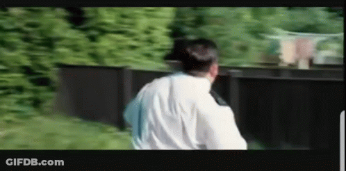 A Man In A White Shirt Is Standing In Front Of A Black Fence . GIF