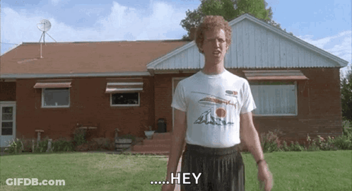 A Man In A White Shirt Is Standing In Front Of A Brick House And Says Hey . GIF