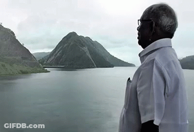 A Man In A White Shirt Is Standing In Front Of A Lake With Mountains In The Background GIF