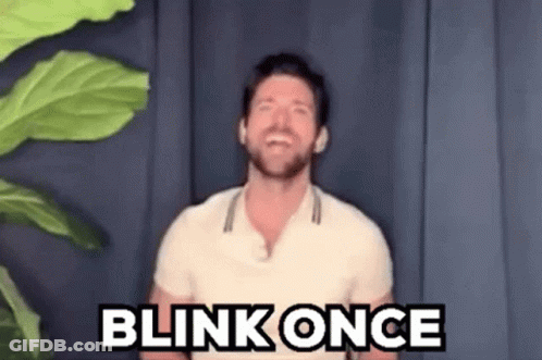 A Man In A White Shirt Is Standing In Front Of A Plant And Saying Blink Once . GIF