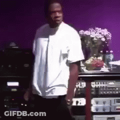 A Man In A White Shirt Is Standing In Front Of A Purple Wall With A Vase Of Flowers In The Background . GIF