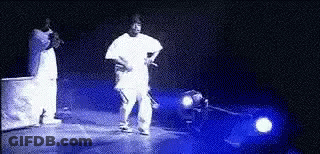 A Man In A White Shirt Is Standing On A Stage In Front Of A Car . GIF