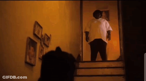 A Man In A White Shirt Is Standing On The Stairs In A Dark Room . GIF