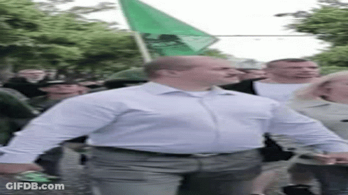 A Man In A White Shirt Is Walking Down A Street With A Green Flag In His Hand . GIF