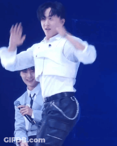 A Man In A White Shirt Is Waving While Another Man Holds A Microphone Behind Him GIF