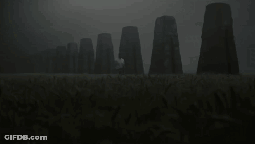 A Man In A White Shirt Stands In A Field With A Row Of Stones In The Background GIF