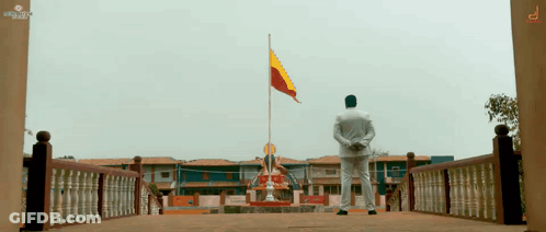 A Man In A White Shirt Stands In Front Of A Building With A Flag Flying In The Background GIF