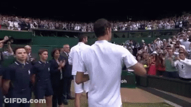 A Man In A White Shirt Stands In Front Of A Crowd Of People On A Tennis Court GIF