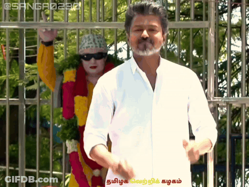 A Man In A White Shirt Stands In Front Of A Statue Of A Woman In A Yellow And Red Outfit GIF