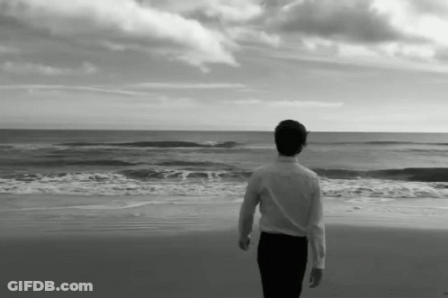 A Man In A White Shirt Stands On A Beach Looking At The Ocean GIF