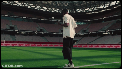 A Man In A White Shirt Stands On A Soccer Field In Front Of A Stadium That Says Stadium Tour GIF