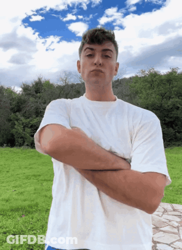 A Man In A White Shirt Stands With His Arms Crossed In A Field GIF