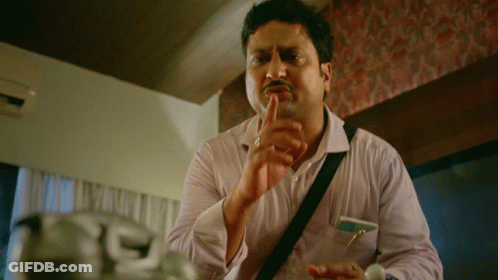 A Man In A White Shirt With A Black Strap Around His Shoulder Holds His Finger To His Lips GIF