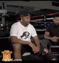 A Man In A White Shirt With A Skull And Crossbones On It Sits Next To Another Man In A Black Shirt GIF