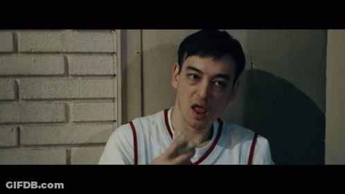 A Man In A White Shirt With Red Stripes On The Sleeves Is Leaning Against A Brick Wall GIF