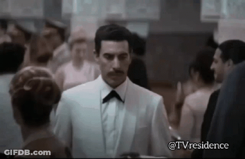 A Man In A White Suit And Black Bow Tie Is Walking Through A Crowd Of People . GIF