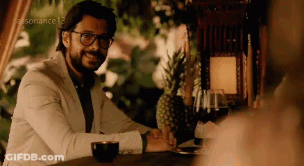A Man In A White Suit And Glasses Is Sitting At A Table . GIF