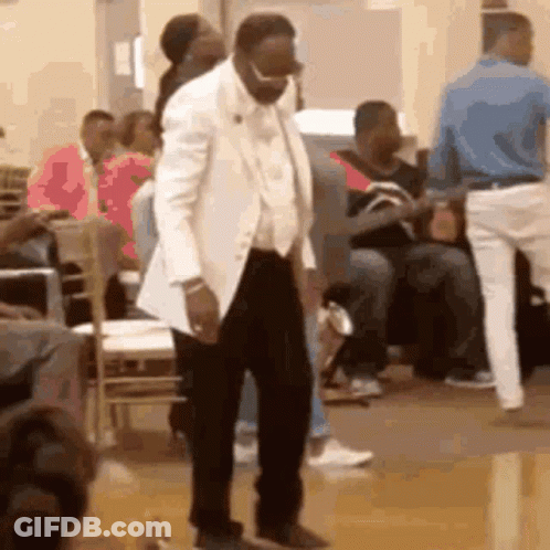 A Man In A White Suit Is Dancing In Front Of A Crowd Of People In A Room . GIF