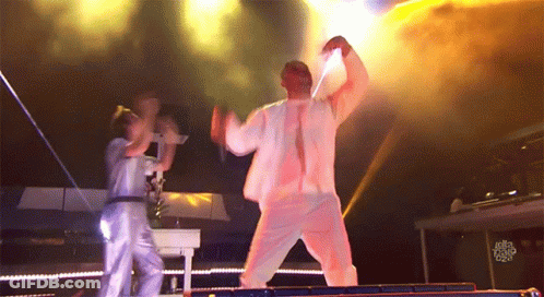 A Man In A White Suit Is Dancing On A Stage While A Woman Stands Behind Him With A Sign That Says Just Dance GIF