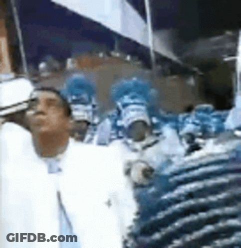 A Man In A White Suit Is Standing In Front Of A Crowd Of People In Blue Costumes . GIF