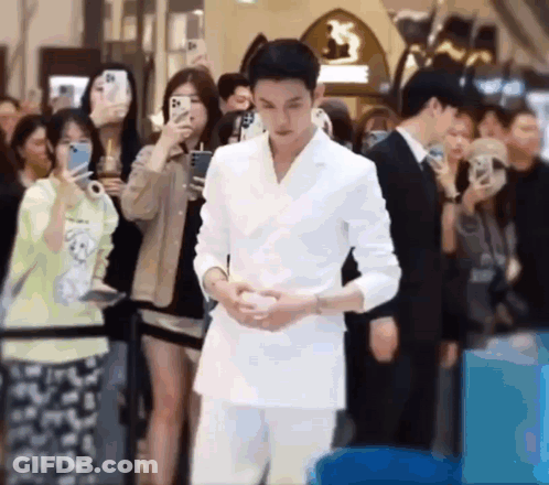 A Man In A White Suit Is Standing In Front Of A Crowd Of People Taking Pictures . GIF