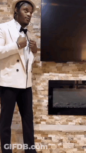 A Man In A White Suit Is Standing In Front Of A Fireplace And Television GIF