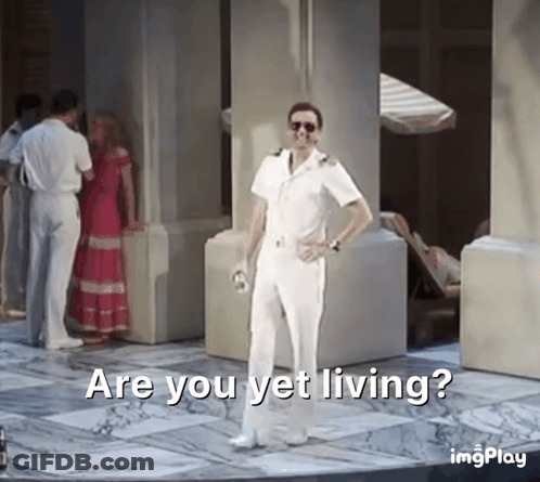 A Man In A White Suit Is Standing In Front Of A Group Of People And Says Are You Yet Living GIF
