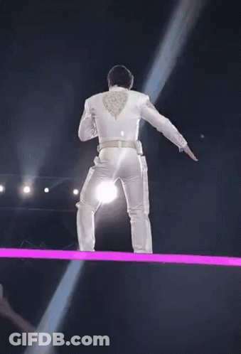 A Man In A White Suit Is Standing On A Stage With A Light Coming From His Back . GIF