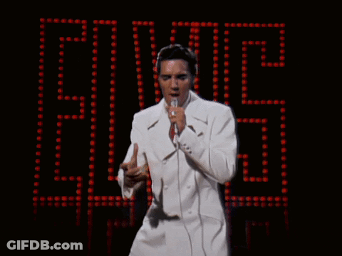 A Man In A White Suit Sings Into A Microphone In Front Of A Elvis Sign GIF