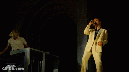 A Man In A White Suit Sings Into A Microphone While Another Man Plays A Keyboard In The Background GIF