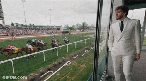 A Man In A White Suit Stands In Front Of A Horse Race GIF