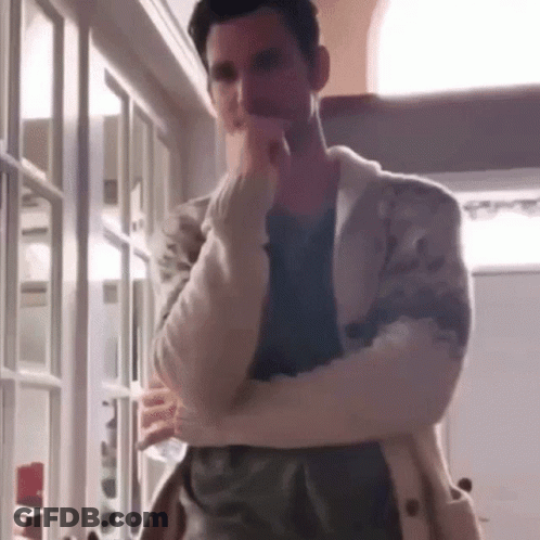 A Man In A White Sweater Is Standing In A Room With His Arms Crossed And His Hand On His Chin . GIF