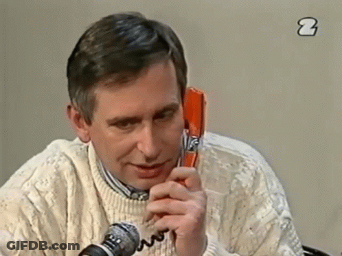 A Man In A White Sweater Is Talking On A Red Telephone GIF