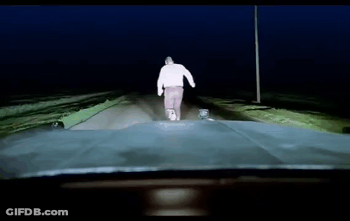 A Man In A White Sweater Is Walking Down A Road At Night GIF