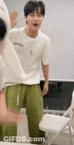 A Man In A White T-Shirt And Green Pants Is Standing Next To A Folding Chair . GIF