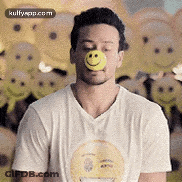 A Man In A White T-Shirt Is Blowing A Yellow Ball With A Smiley Face On His Nose . GIF