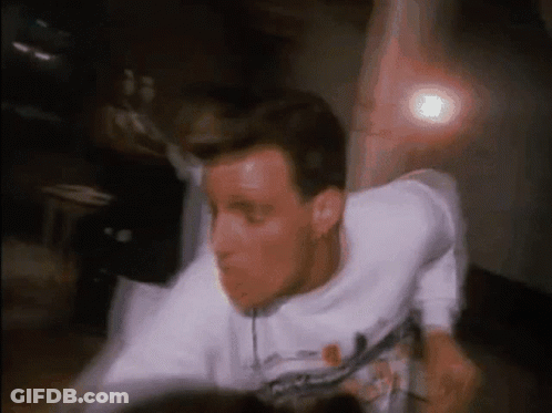 A Man In A White T-Shirt Is Dancing In Front Of A Light GIF