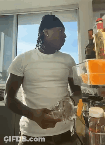 A Man In A White T-Shirt Is Standing In Front Of A Fridge Filled With Bottles And Condiments GIF