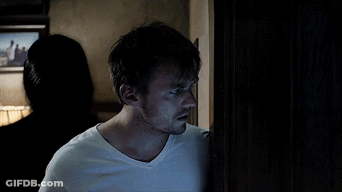 A Man In A White T-Shirt Stands In A Dark Room GIF
