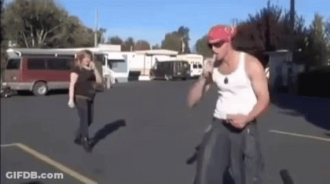 A Man In A White Tank Top And A Red Bandana Is Standing In A Parking Lot With A Woman Walking Behind Him . GIF
